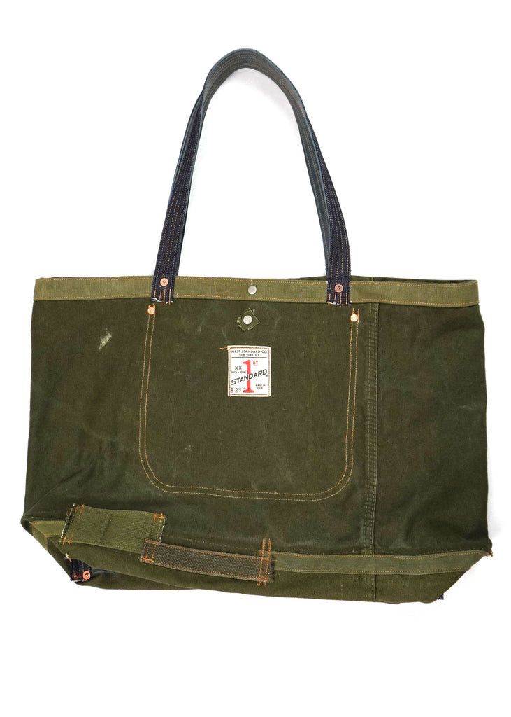 The First Standard Duffle Tote Bag 82924 is a durable and versatile carryall crafted with high-quality materials. Designed in a rugged olive-green waxed canvas, it features contrast stitching and reinforced denim handles for comfortable and sturdy handling. The exterior is adorned with a First Standard Co. logo patch for a vintage-inspired aesthetic.

A front pocket with secure stitching adds extra utility, while copper rivet accents ensure longevity. 